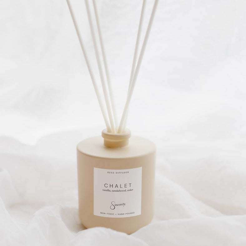A beige reed diffuser bottle with six white reeds, labeled 'CHALET', set against a soft white draped fabric background.