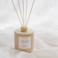 A reed diffuser bottle in a warm beige tone with six natural reed sticks, resting on draped white fabric. The label reads 'GOLDEN' 