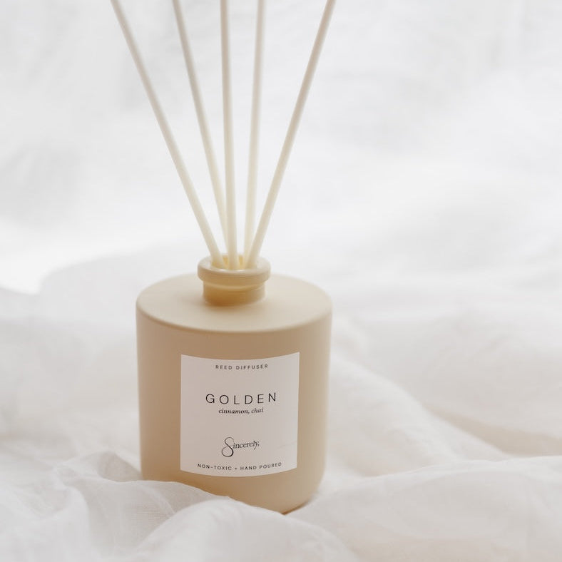 A reed diffuser bottle in a warm beige tone with six natural reed sticks, resting on draped white fabric. The label reads 'GOLDEN' 