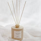 A reed diffuser bottle in a warm beige tone with five natural reed sticks, set against a soft white draped fabric background. The label reads 'GOLDEN'.