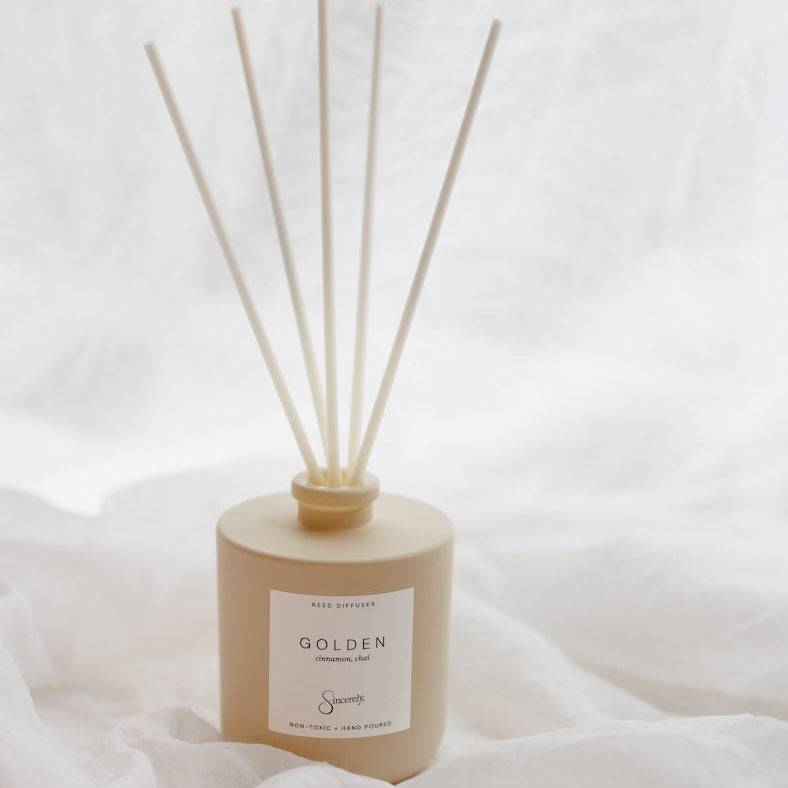 A reed diffuser bottle in a warm beige tone with five natural reed sticks, set against a soft white draped fabric background. The label reads 'GOLDEN'.