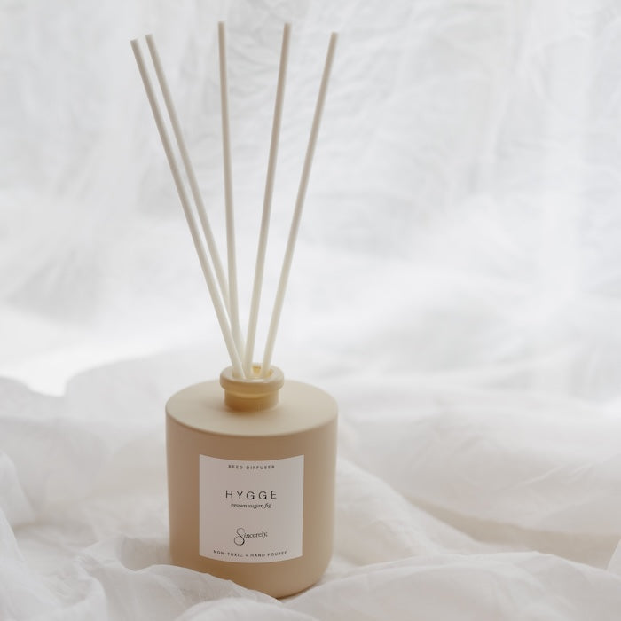 A reed diffuser bottle in a matte beige finish with six natural wood reeds, resting on draped white fabric.