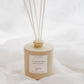 A beige reed diffuser bottle with six natural reed sticks, resting on white draped fabric. The bottle features a white label with the scent 'FLOURISH'.