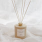 A reed diffuser bottle in a matte beige finish with six natural reed sticks, set against a soft white draped fabric background. The label reads 'AUTUMN LEAVES'