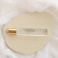 A clear glass perfume roller bottle with a gold cap, labeled 'PARFUM ROLLER FLOURISH', resting on a textured beige oval pad over a soft white fabric background.