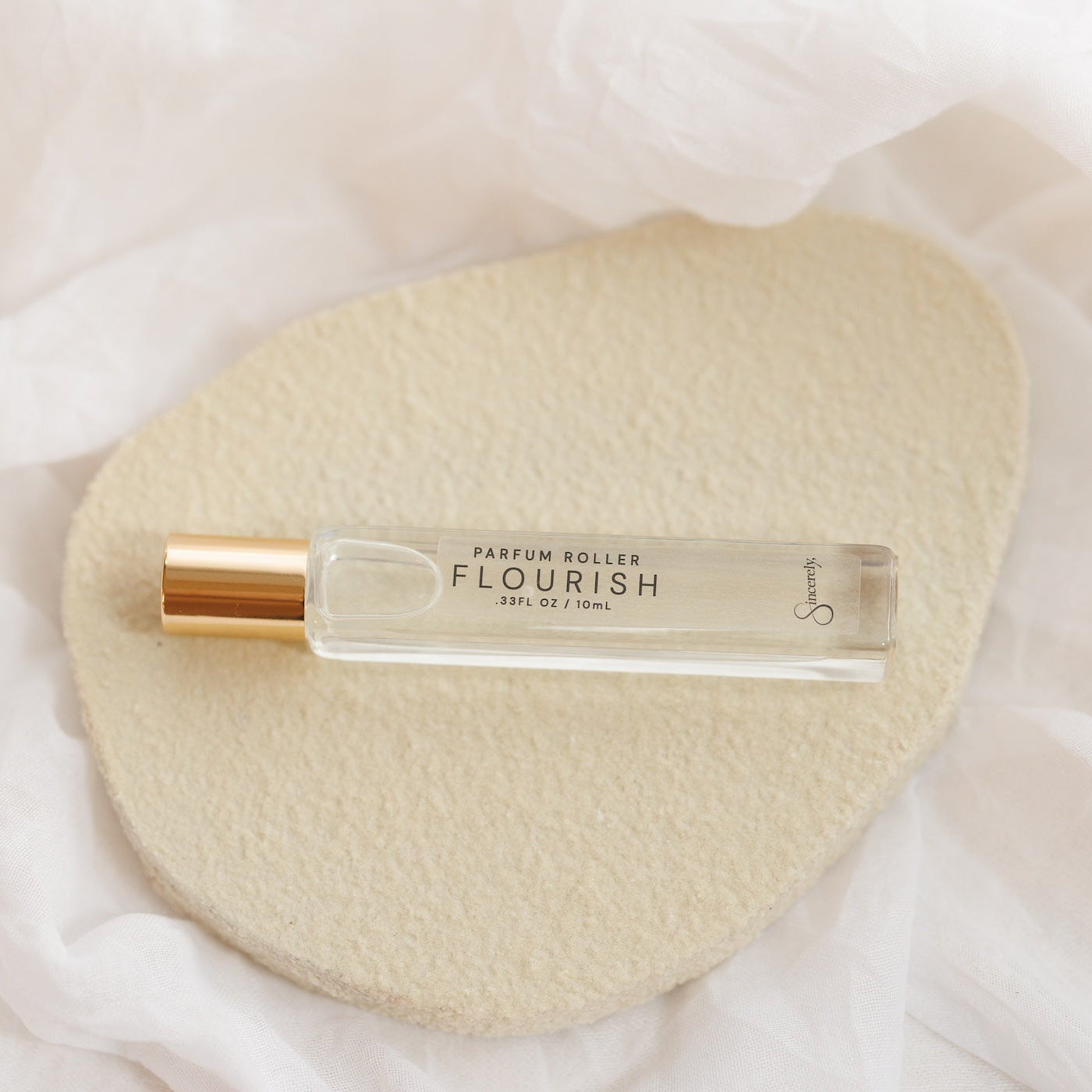 A clear glass perfume roller bottle with a gold cap, labeled 'PARFUM ROLLER FLOURISH', resting on a textured beige oval pad over a soft white fabric background.