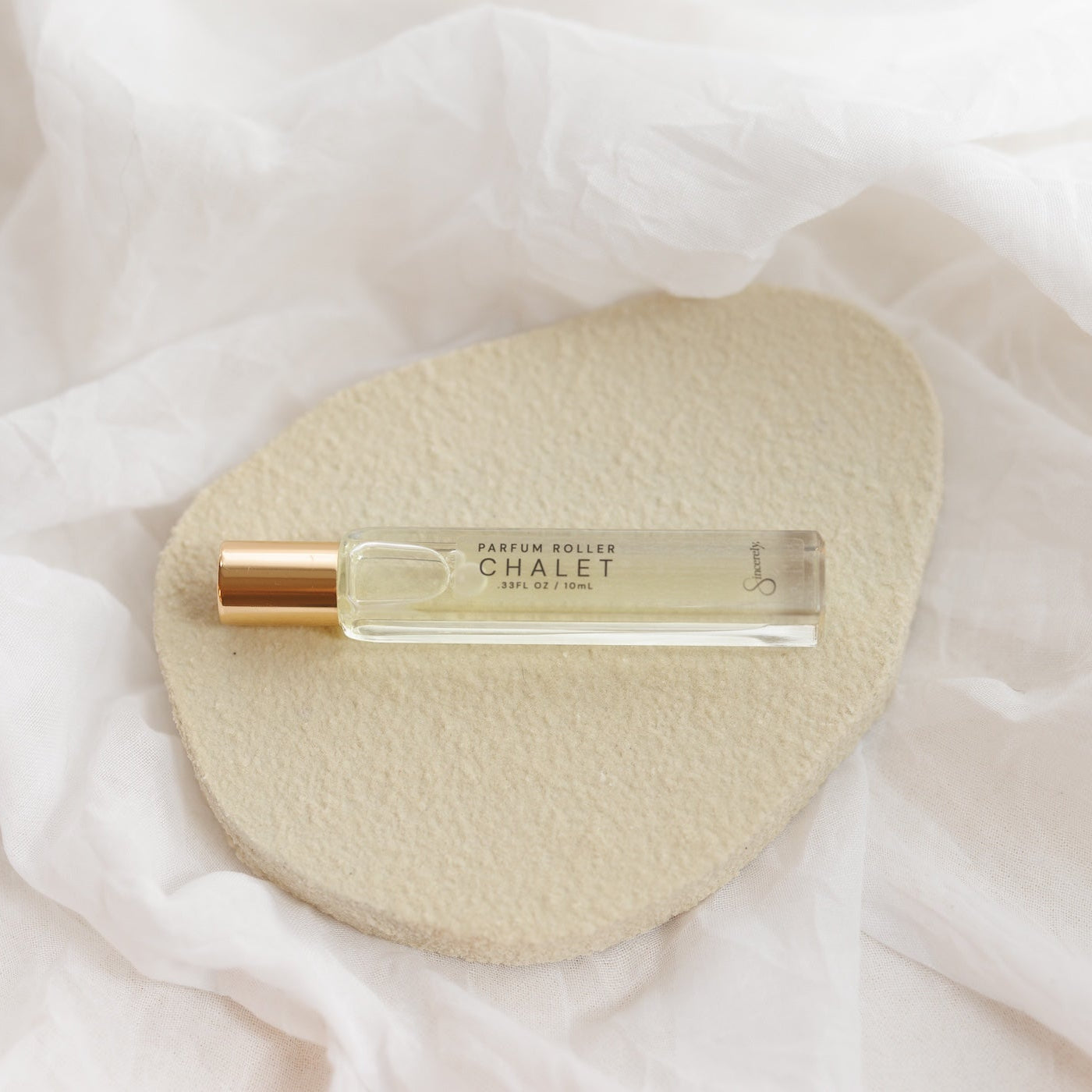 A small clear glass perfume roller bottle with a gold cap, labeled CHALET, resting on a textured beige stone over a soft white fabric background.