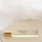 A small clear glass perfume roller bottle with a metallic gold cap, labeled 'CHALET', resting on a textured beige stone against a soft white draped fabric background.