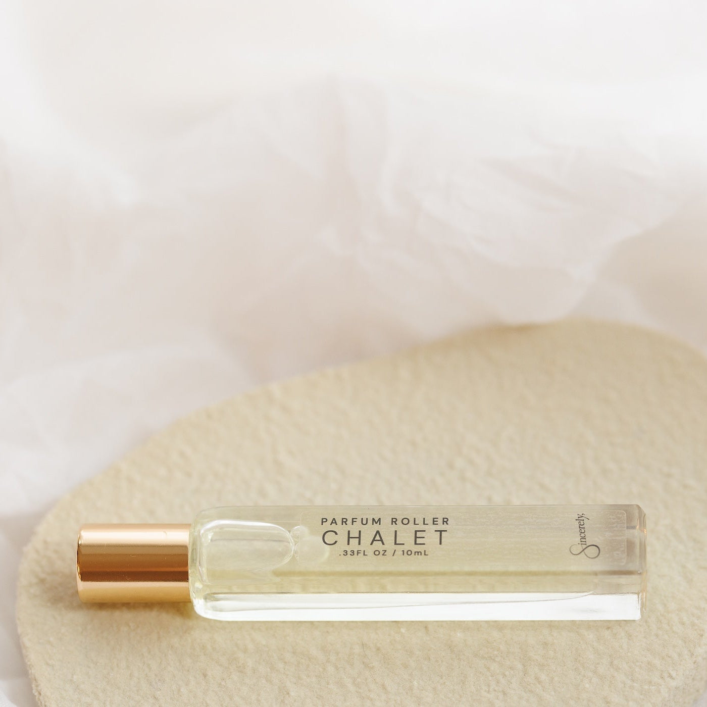 A small clear glass perfume roller bottle with a metallic gold cap, labeled 'CHALET', resting on a textured beige stone against a soft white draped fabric background.
