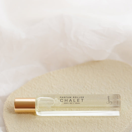 A small clear glass perfume roller bottle with a metallic gold cap, labeled 'CHALET', resting on a textured beige stone against a soft white draped fabric background.