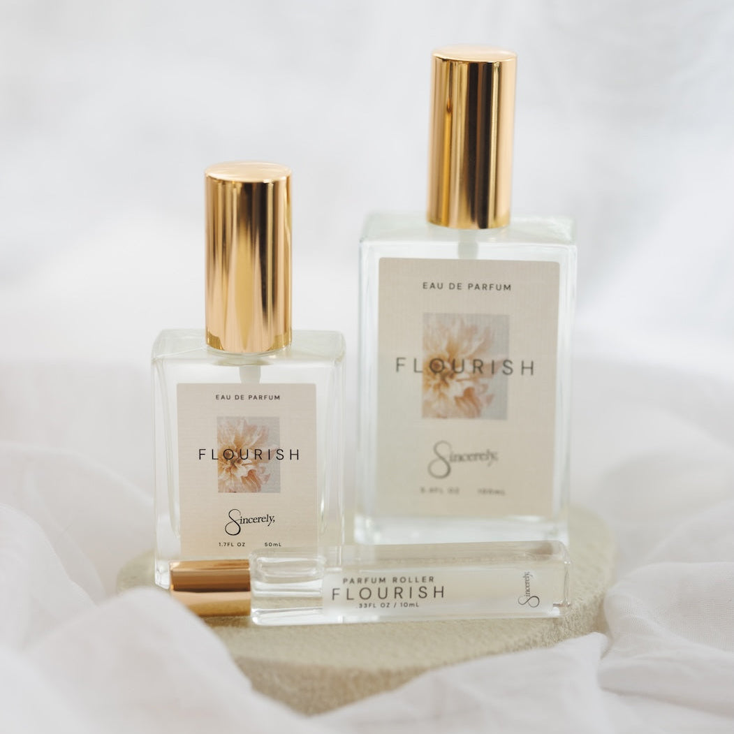 Two clear glass perfume bottles with gold caps and cream labels reading FLOURISH, displayed on a light stone base with draped white fabric around them.
