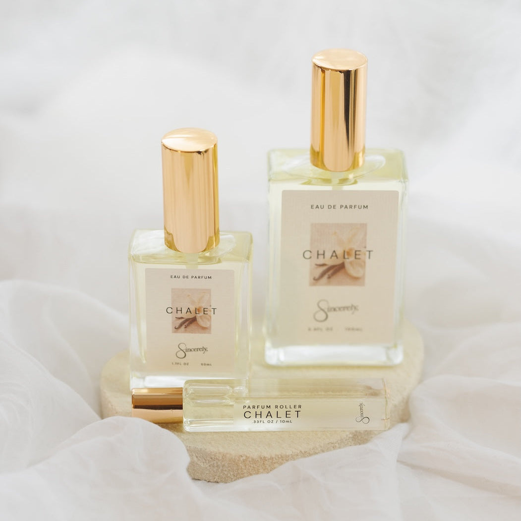 Two CHALET Eau de Parfum bottles with gold caps and a small matching parfum roller, displayed on a wooden block with white draped fabric in the background.
