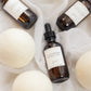 Three amber glass dropper bottles with white labels are arranged on white fabric with large white felt balls around them. The center bottle label reads FLOURISH.