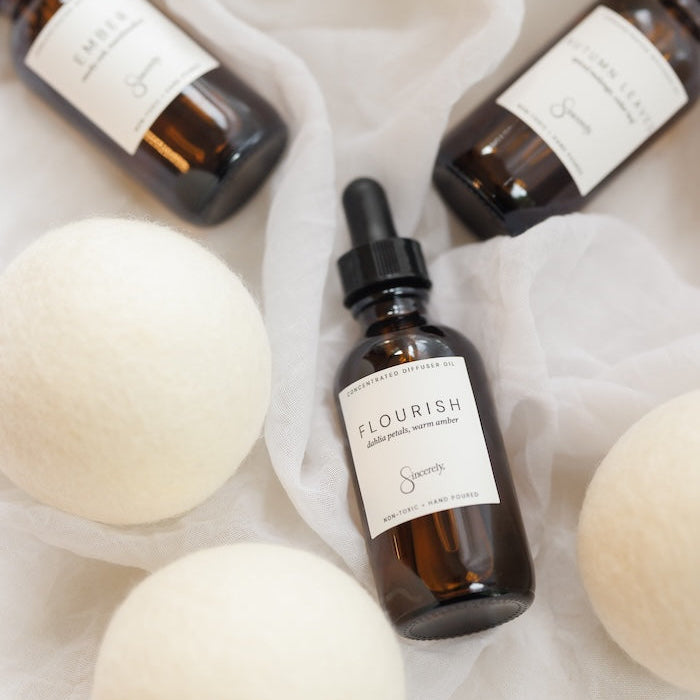 Three amber glass dropper bottles with white labels are arranged on white fabric with large white felt balls around them. The center bottle label reads FLOURISH.
