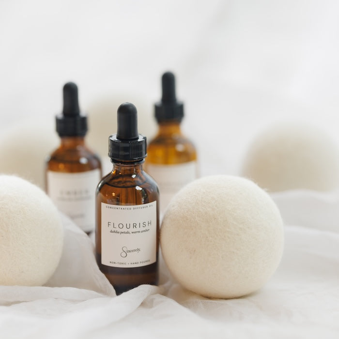 Three amber glass dropper bottles with black caps and white labels, arranged on white fabric with soft white felt balls around them. The front bottle label reads FLOURISH.