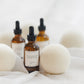 Three amber glass dropper bottles with black caps and white labels, arranged on white fabric with soft white felt balls around them. The front bottle label reads FLOURISH.