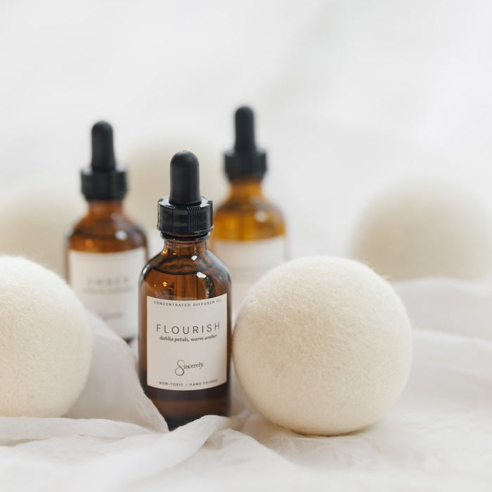 Three amber glass dropper bottles with black caps and white labels, arranged on white fabric with soft white felt balls around them. The front bottle label reads FLOURISH.