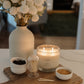 A lit scented candle in a clear glass jar sits on a wooden board with small white bowls, a match container, and a large vase of white flowers on a tabletop in a softly blurred indoor setting.