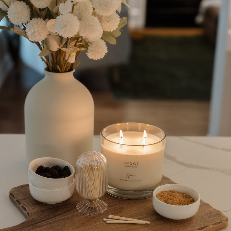 A lit scented candle in a clear glass jar sits on a wooden board with small white bowls, a match container, and a large vase of white flowers on a tabletop in a softly blurred indoor setting.