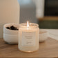 A lit soy candle in a clear glass jar sits on a wooden surface in a softly blurred indoor setting. The label reads 'HYGGE'.
