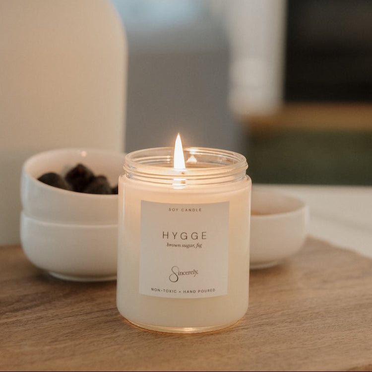 A lit soy candle in a clear glass jar sits on a wooden surface in a softly blurred indoor setting. The label reads 'HYGGE'.