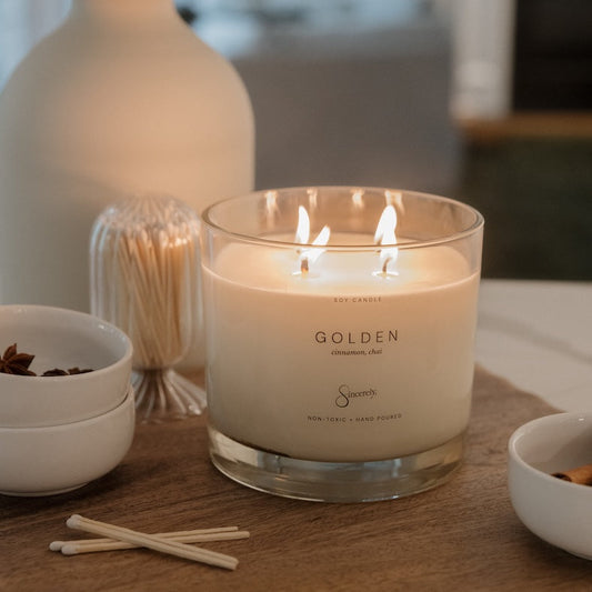 A lit 4-wick candle in a clear glass jar sits on a wooden table, surrounded by dried flowers in a white vase, small white bowls, and matches. The candle label reads 'GOLDEN'.