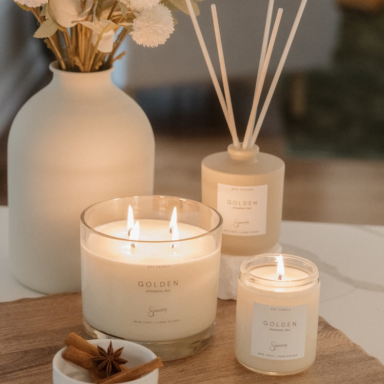 A styled home scene featuring a set of Golden scented home fragrance products, including a large 4-wick candle, a smaller jar candle, and a reed diffuser, arranged on a wooden cutting board next to a white vase with dried flowers.