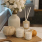 A styled home decor arrangement featuring three 'Harvest' scented candles in glass jars, a reed diffuser, and a large vase of white flowers, all on a wooden board with small pumpkins on a white marble surface.