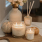 A styled home decor scene featuring a large white candle in a glass jar on a wooden board, a smaller matching candle, a reed diffuser, a ceramic vase with white flowers, and two small pumpkins on a white tabletop.