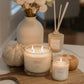 A styled home decor scene featuring a large white candle in a glass jar on a wooden board, a smaller matching candle, a reed diffuser, a ceramic vase with white flowers, and two small pumpkins on a white tabletop.