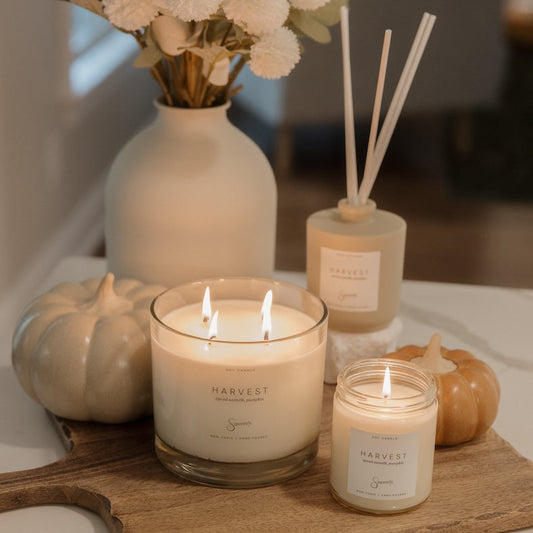 A styled home decor scene featuring a large white candle in a glass jar on a wooden board, a smaller matching candle, a reed diffuser, a ceramic vase with white flowers, and two small pumpkins on a white tabletop.