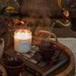 A cozy lifestyle scene featuring a lit scented candle labeled 'EMBER' on a woven tray, alongside dark brown ceramic teacups, a book, and steam rising from a teapot pouring into one cup.