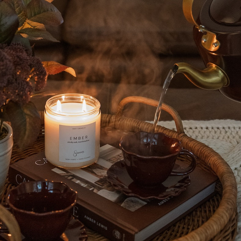 A cozy lifestyle scene featuring a lit scented candle labeled 'EMBER' on a woven tray, alongside dark brown ceramic teacups, a book, and steam rising from a teapot pouring into one cup.