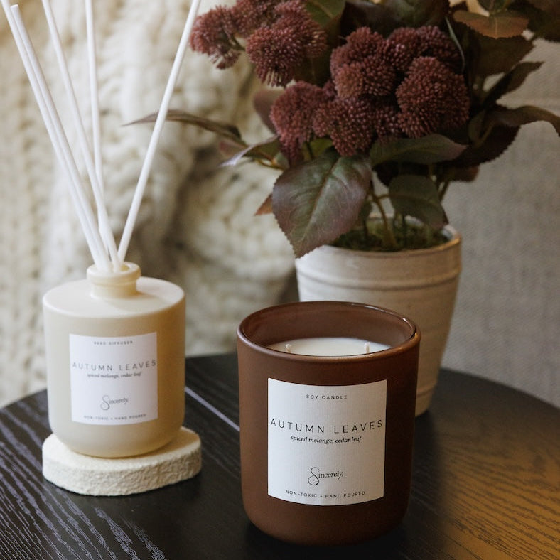 A cozy home setting featuring a brown glass candle jar and a beige reed diffuser bottle with white reeds, both labeled 'Autumn Leaves', styled on a dark wooden table next to a potted plant.