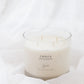 A 4-wick soy candle in a clear glass jar with cream-colored wax, labeled 'EMBER', set against a soft white draped fabric background.
