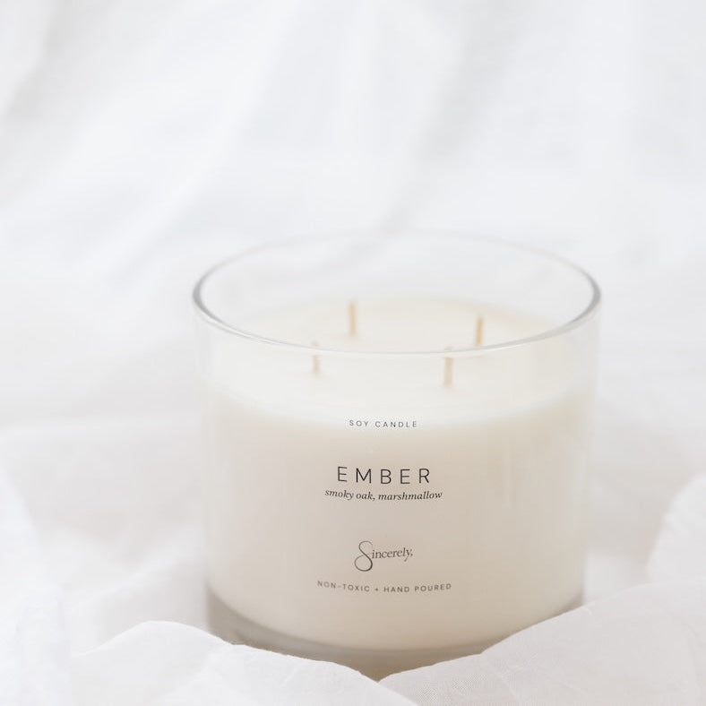 A 4-wick soy candle in a clear glass jar with cream-colored wax, labeled 'EMBER', set against a soft white draped fabric background.
