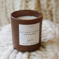 A brown ceramic soy candle jar labeled 'Autumn Leaves', resting on a chunky white knit blanket with a matching knit background.