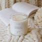 A soy candle in a clear glass jar with a white label reading 'CHALET', styled on a chunky white knit blanket with an open book and pale flowers in the background.