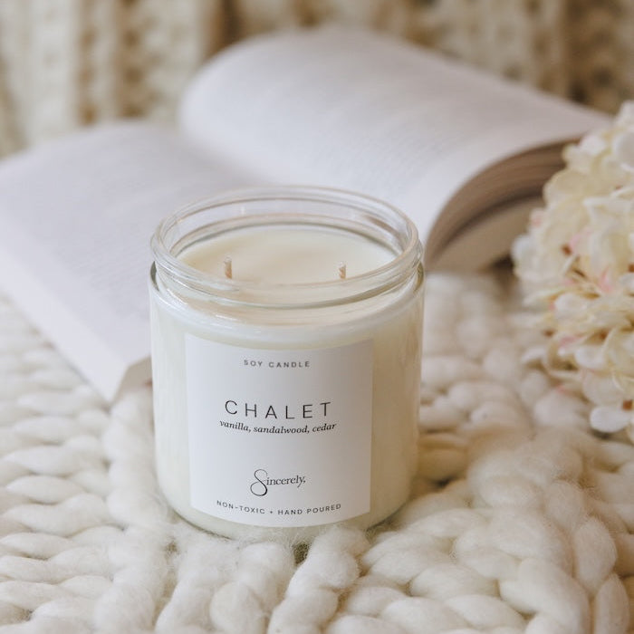A soy candle in a clear glass jar with a white label reading 'CHALET', styled on a chunky white knit blanket with an open book and pale flowers in the background.