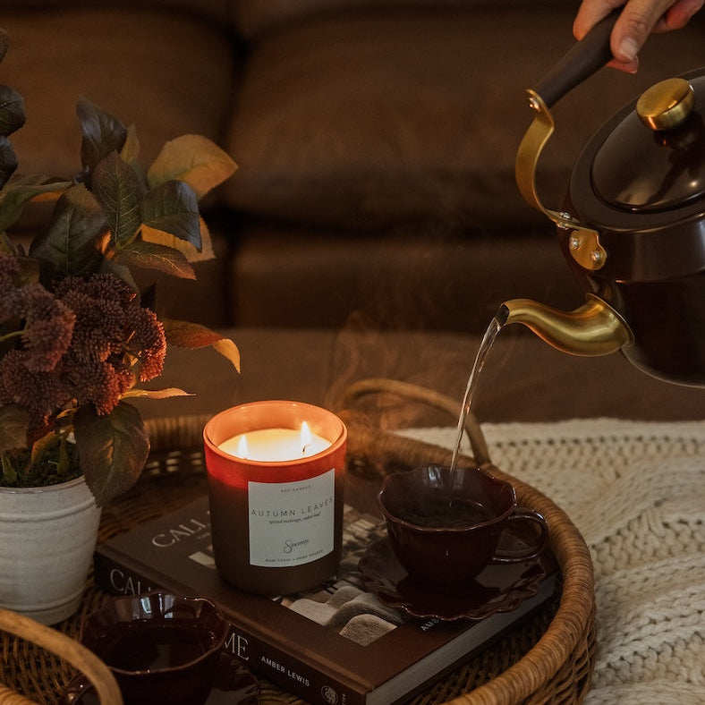 A cozy lifestyle scene showing a lit scented candle on a wicker tray, with a hand pouring hot water from a brass kettle into a dark cup. A wicker candle tin and a potted plant are also arranged on the tray.