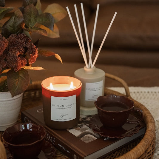 A cozy autumn-themed home decor arrangement featuring a lit candle, a reed diffuser, and a book on a woven tray, styled with dark ceramic cups and a potted plant in a warm living room setting.