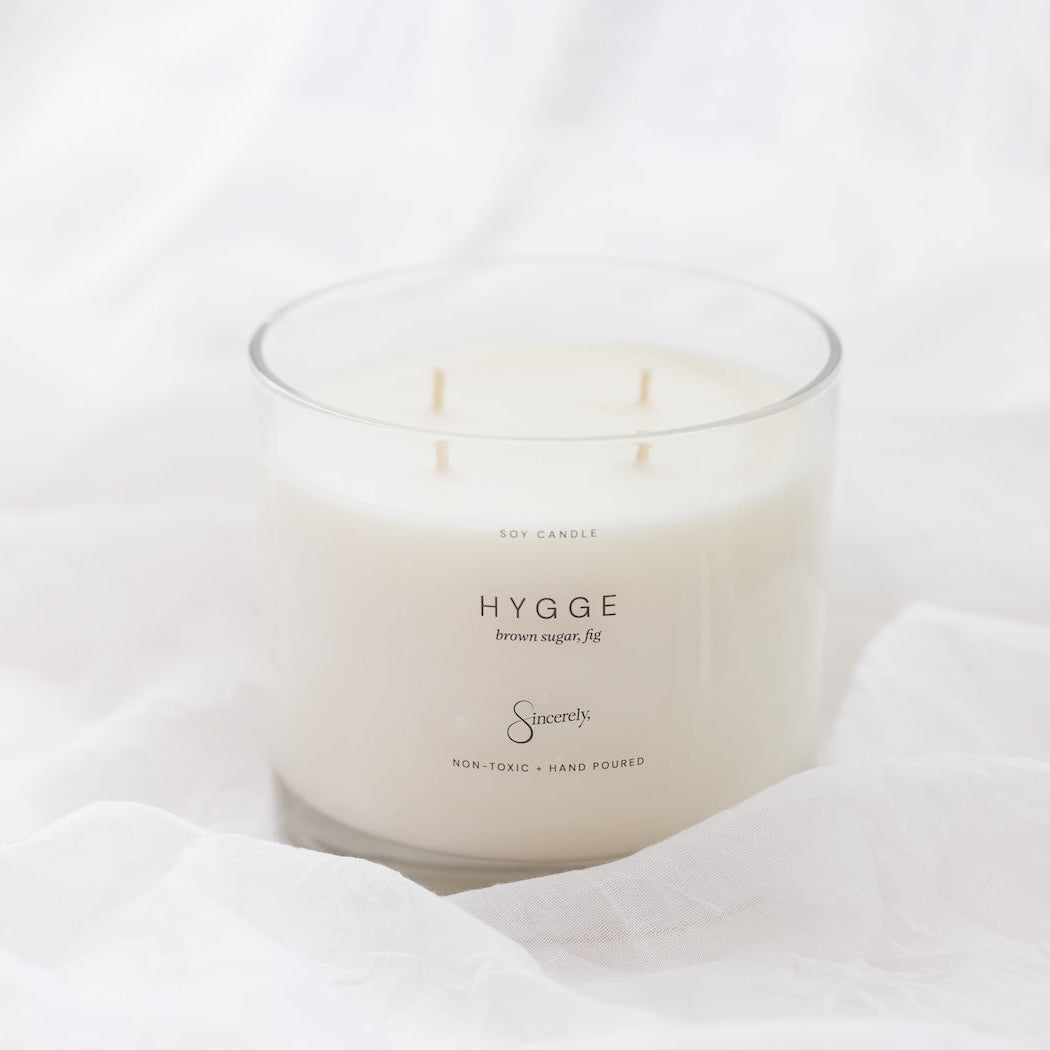 A 4-wick soy candle in a clear glass jar with creamy off-white wax, resting on draped white fabric. The label reads 'HYGGE'