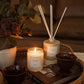 A cozy indoor scene showing a lit candle and a reed diffuser bottle with reeds, arranged on a wicker tray with books and dark bowls. The labels on both products read HYGGE.