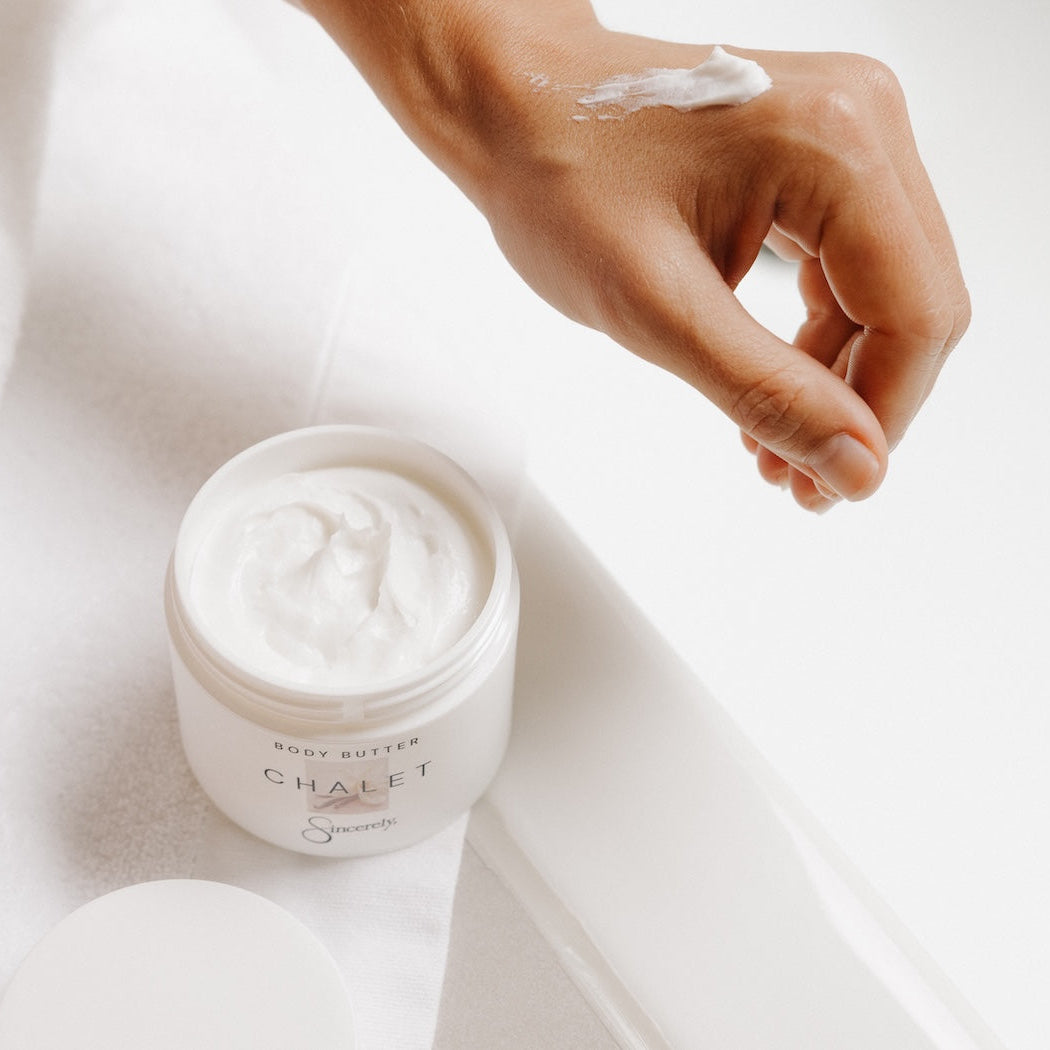 An open white jar of Chalet body butter with white cream visible inside, placed on a white towel beside a person's hand that has a dollop of the cream on it.