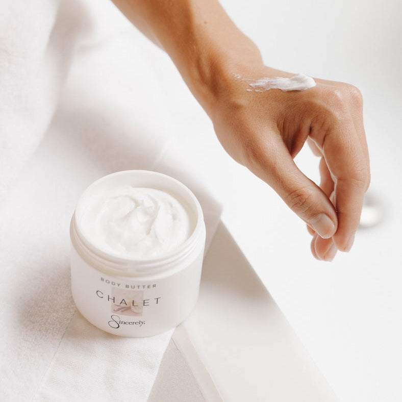 A person applies white body butter cream from a white jar labeled CHALET  onto their hand, set against a clean white background with a white towel.