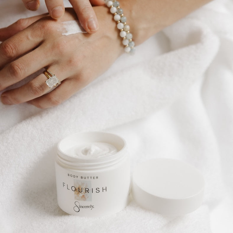 An open white jar of body butter labeled 'FLOURISH' sits on a white towel, with a person's hands applying white cream to one hand. The person is also wearing a pearl beaded bracelet and a gold ring.