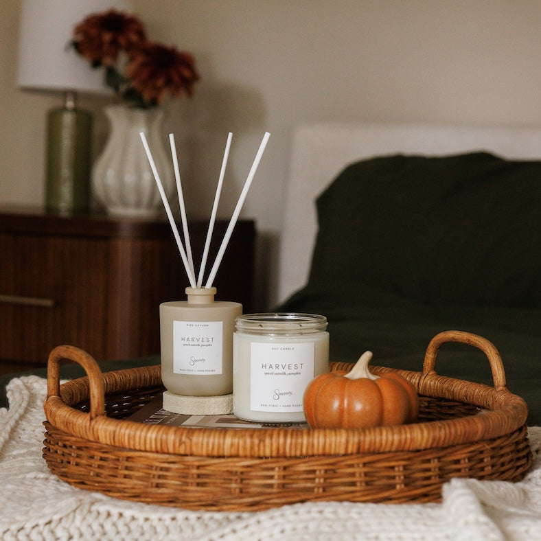 A styled bedroom scene showing a wicker tray holding a reed diffuser, a candle jar labeled HARVEST, and a small orange pumpkin on a bed with a knit blanket.