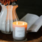 A lit soy candle in a frosted glass jar sits on a woven tray, with a white vase of flowers and an open book softly blurred in the background.