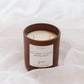 A brown glass jar candle with cream-colored wax and a white label reading 'Autumn Leaves'  styled on white draped fabric.