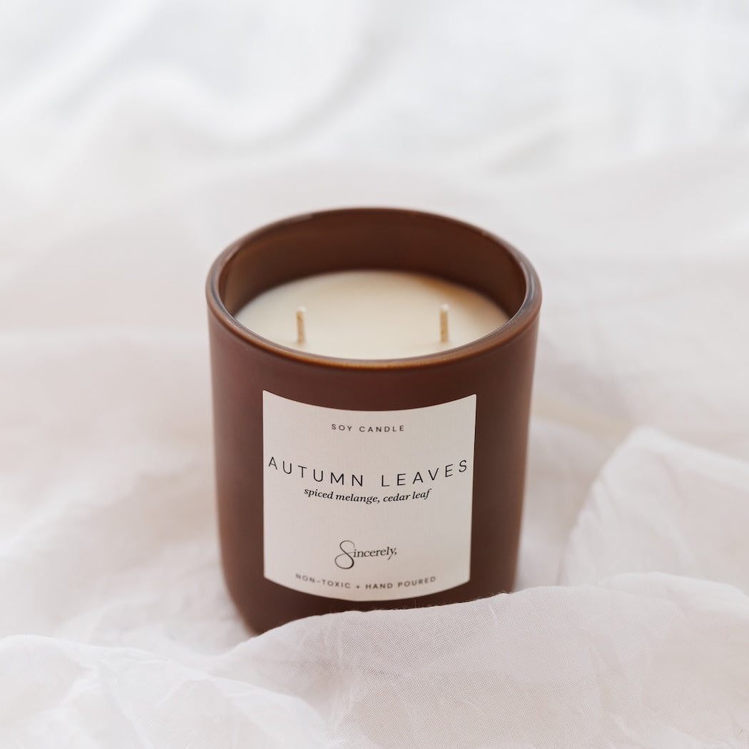 A brown glass jar candle with cream-colored wax and a white label reading 'Autumn Leaves'  styled on white draped fabric.